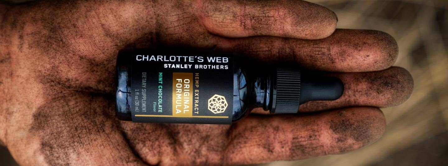 An open hand covered in soil with a bottle of Charlotte's Web CBD Oil in their palm.