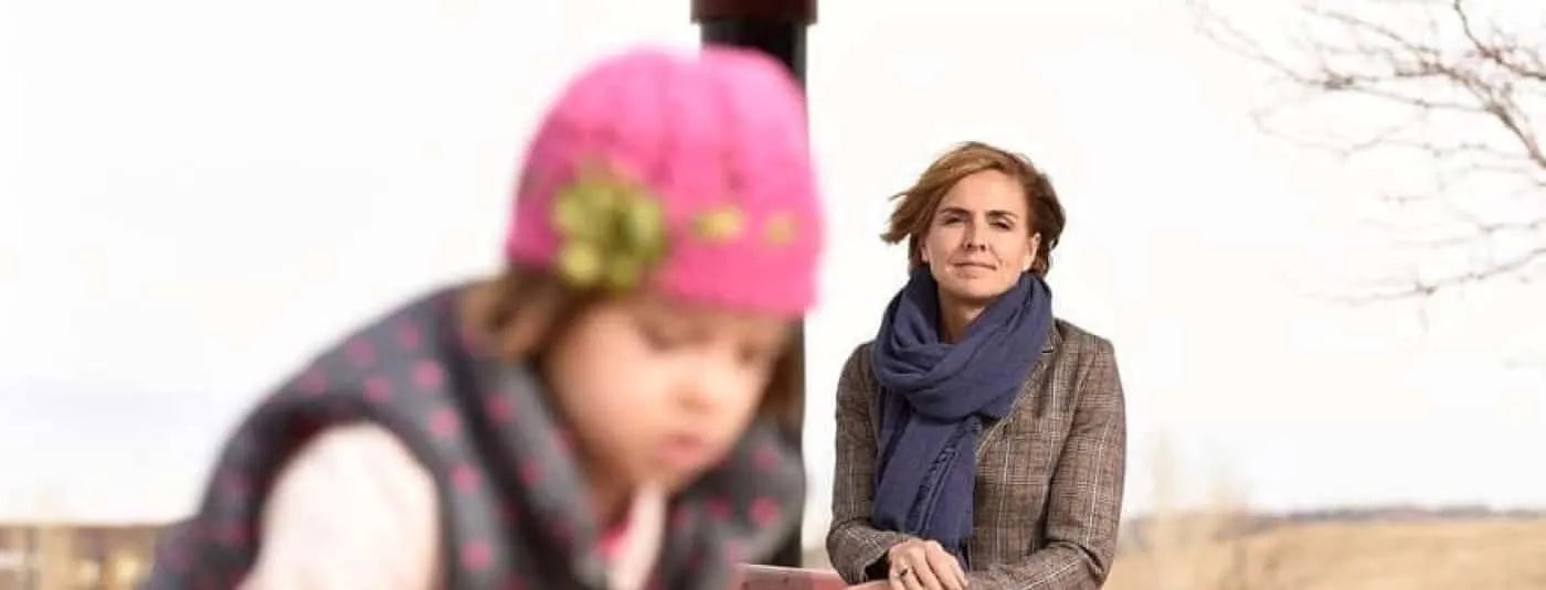 A mother looking from the background at her daughter in a pink hat in the foreground.