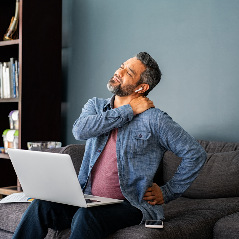 Man sitting on a couch with a laptop, holding his neck and back in discomfort.