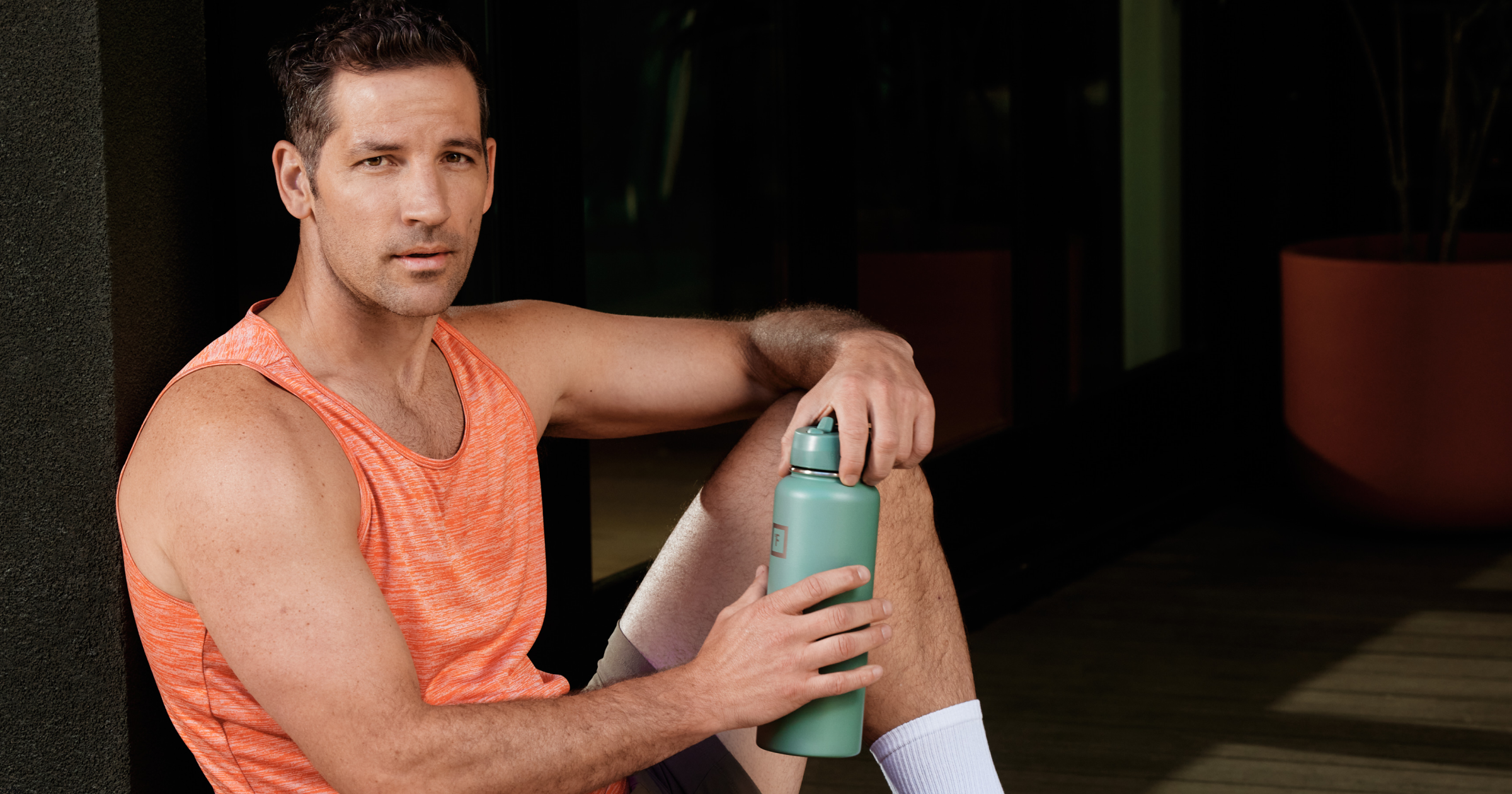 Man in orange tank top leaning against a wall with a water bottle in hand
