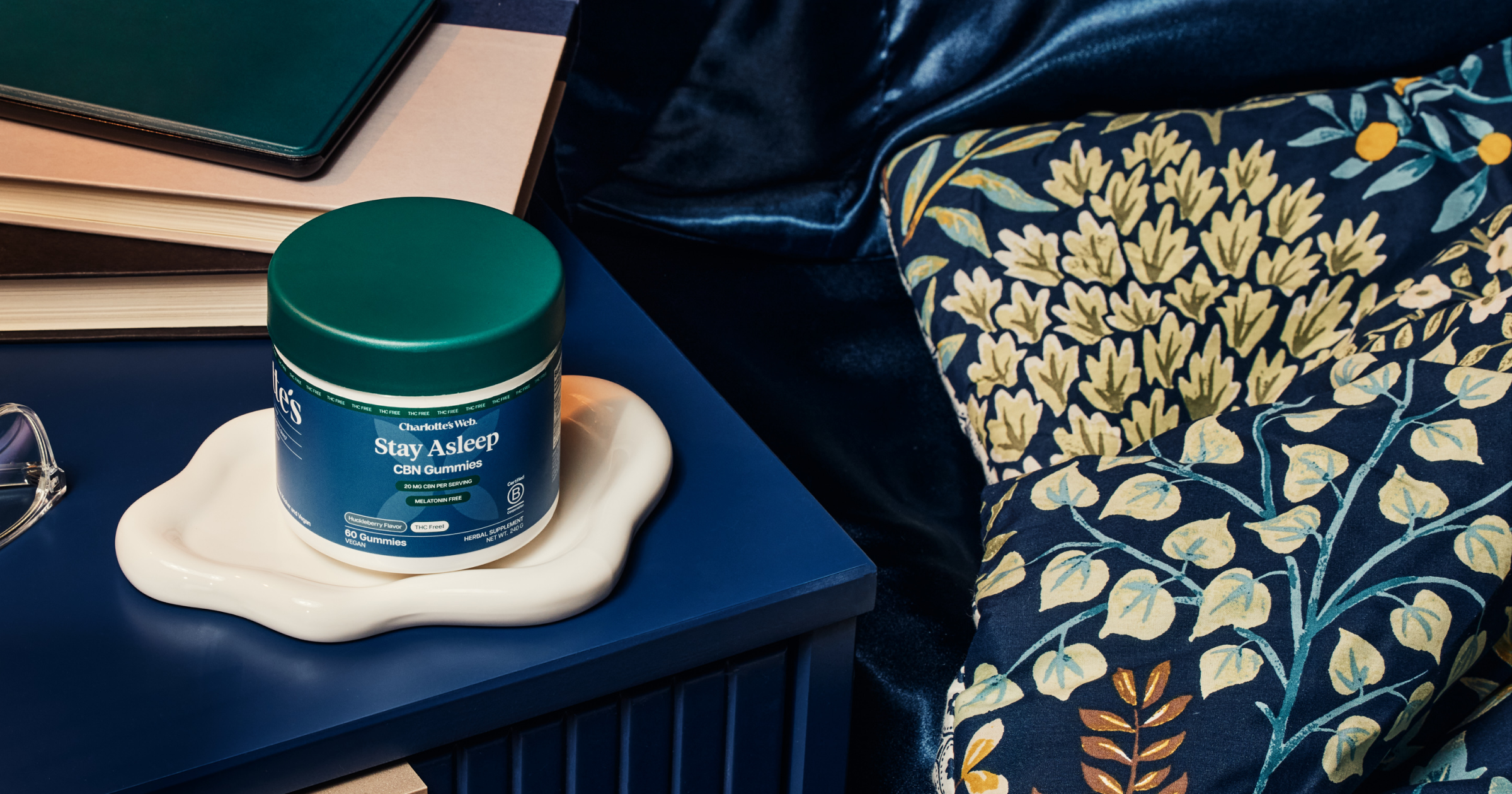 Jar of 'Stay Asleep' supplement on a nightstand next to books and a patterned pillow.
