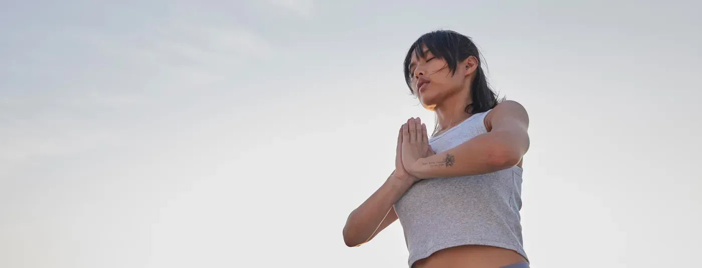 A woman with her eyes closed and her palms together against a cloudy sky.