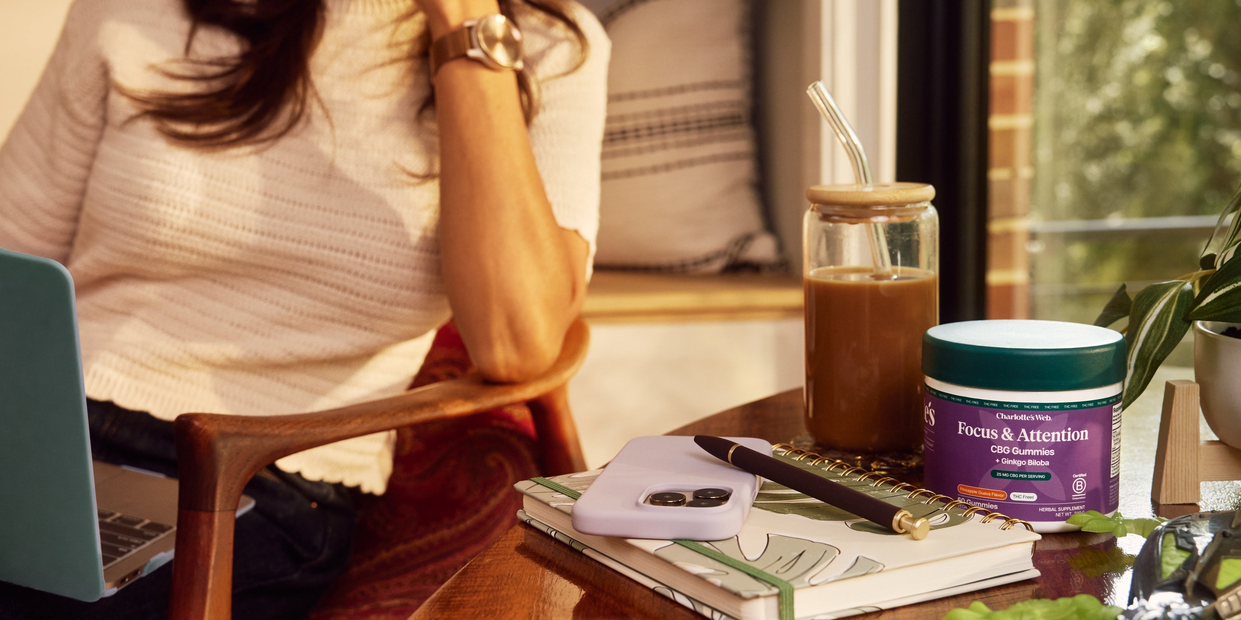 A jar of Charlotte’s Web Focus & Attention CBG Gummies placed on a wooden table beside an iced coffee, notebook, pen, and smartphone, with a woman working on a laptop in the background.