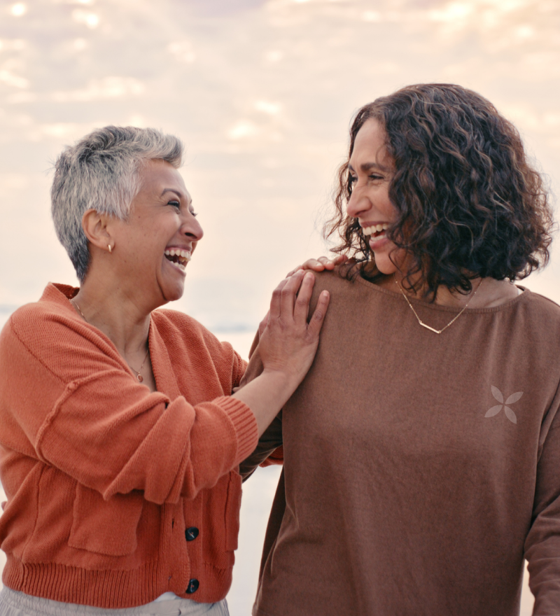 Two women laughing together outdoors with a soft, blurred background