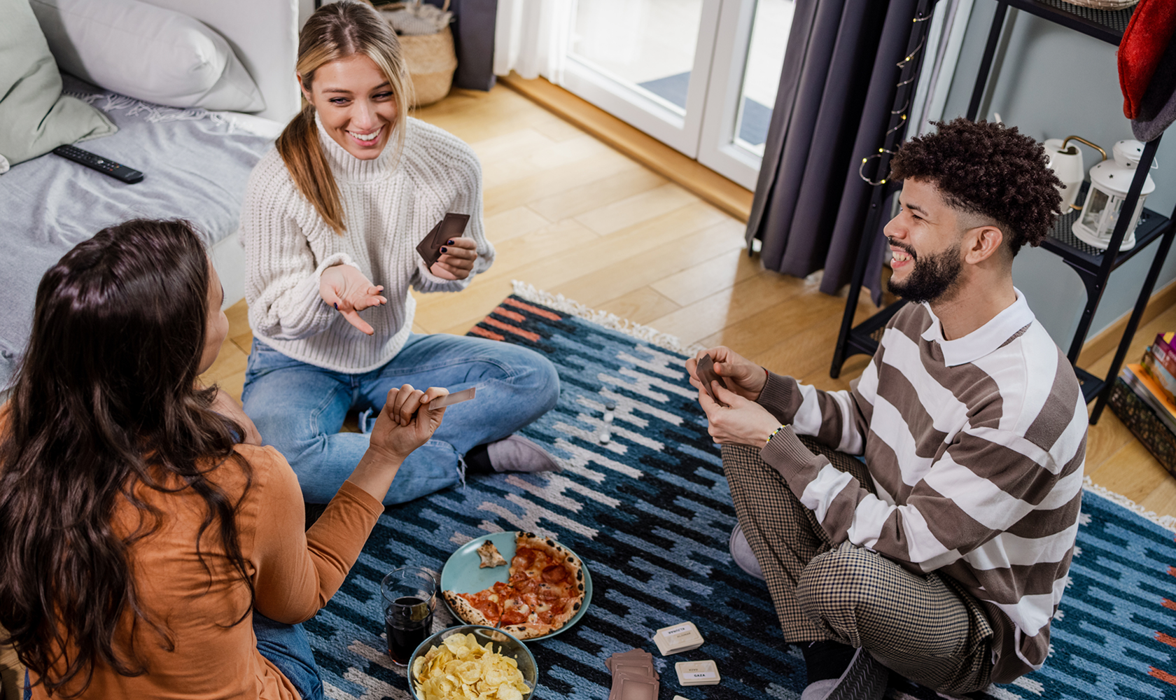 Three people sitting on a rug in a living room, eating snacks and playing cards.
