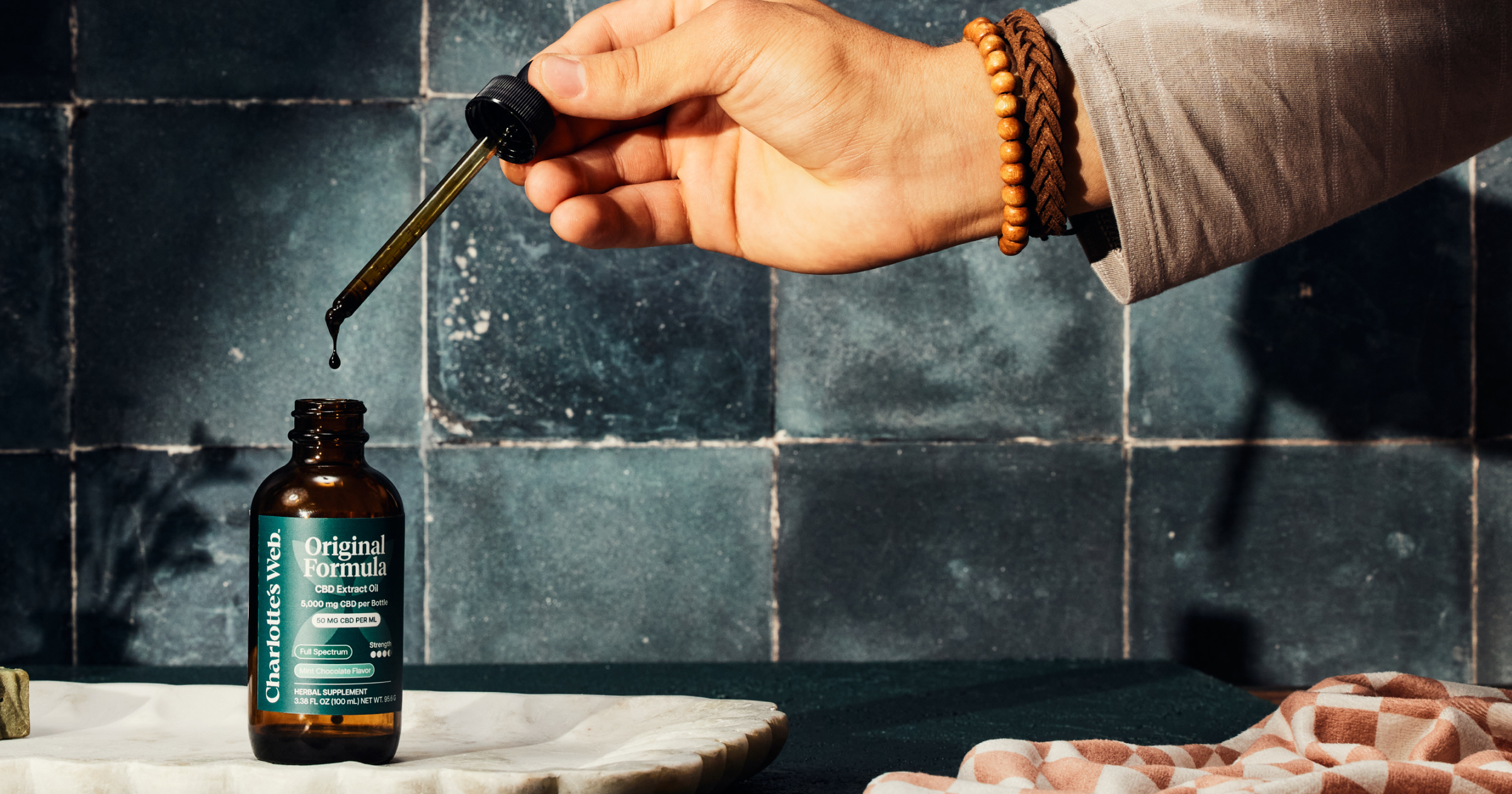 Hand holding a dropper over a bottle labeled 'Original Formula' against a dark tiled wall.