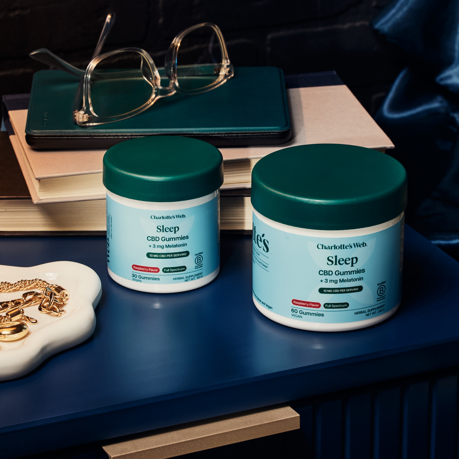 Product Image for Two jars of Charlotte&#39;s Web Sleep CBD Gummies on a nightstand with books and glasses.