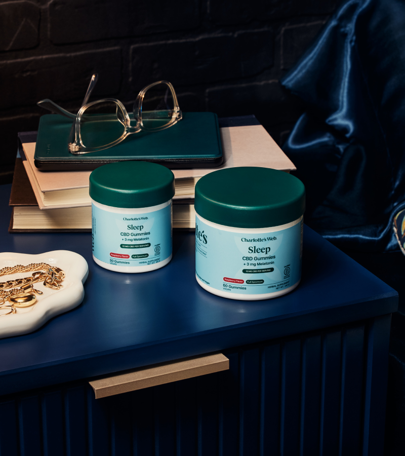 Two jars of 'Sleep' CBD Gummies on a dark surface with books and glasses in the background.