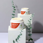 Product Image for Two jars of CBD Clinic Pain Relief Ointment on white stands with green leaves on a gray background